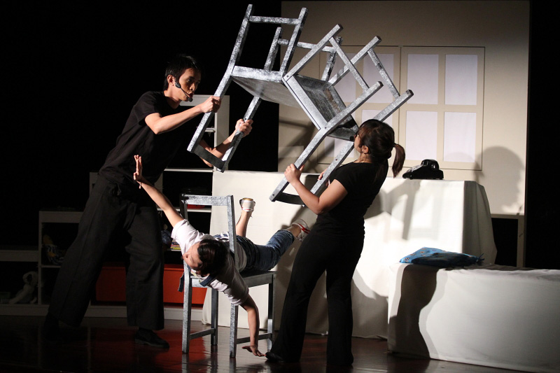 A child playing on a chair with two other actors holding chairs above her in Monster Under My Bed by Paper Monkey