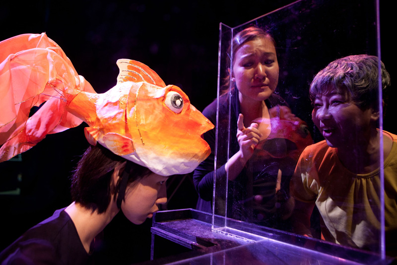 A daughter with an old mother looking through a glass at a gold fish puppet in Goldfish, Go Fish by Paper Monkey