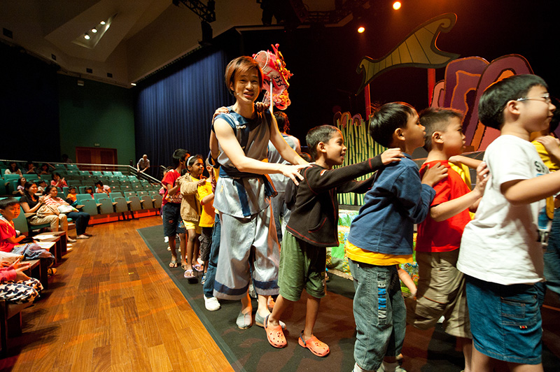 Actor interacting with children audience holding onto each others shoulders Dragon Dance by Paper Monkey Theatre Singapore