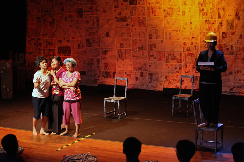 Four housewives chatting happily and a person standing on a chair with a safety helmet on stage left