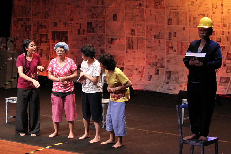 Four housewives gathered in discussion and a person standing on a chair with a safety helmet on stage left