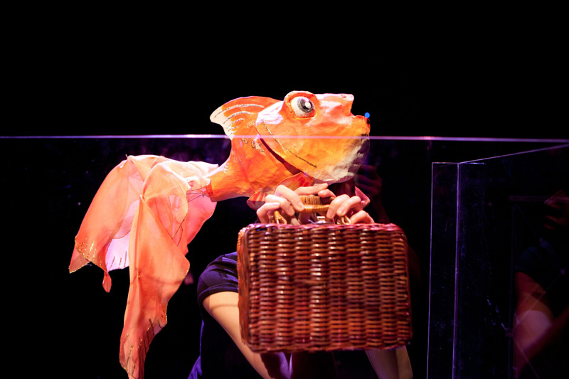 A puppet gold fish holding a basket luggage in Goldfish, Go Fish by Paper Monkey
