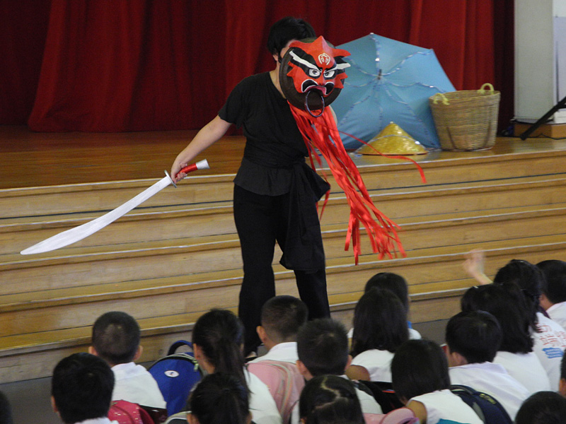 An actor with red mask with a sword prop in front of student audience in Dragon Dance Community Tour by Paper Monkey